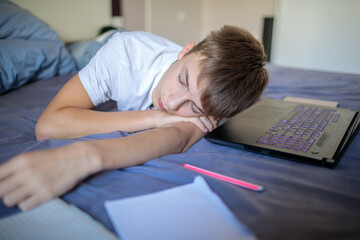 Close-up of a tired or exhausted teenage boy sleeping with his head on his arm next to a laptop after a long session of work or studying