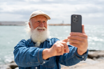 A cheerful elderly man with a white beard, wearing a beige cap and denim shirt, smiles while taking a selfie with his smartphone near the ocean on a sunny day