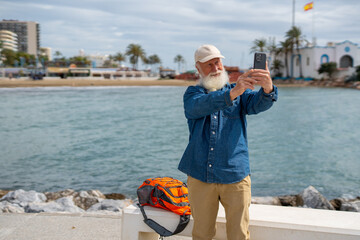 An elderly man with a white beard, wearing a beige cap and denim shirt, takes a selfie with his smartphone by the waterfront, with a Spanish flag, palm trees, and a coastal town in the background
