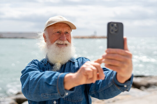 A cheerful elderly man with a white beard, wearing a beige cap and denim shirt, smiles while taking a selfie with his smartphone near the ocean on a sunny day