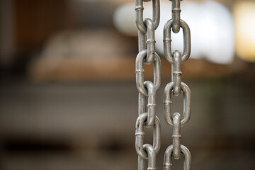 A chain of silver metal links is hanging from a chain link fence. The chain is long and thin, and the links are spaced evenly apart. The image has a somewhat industrial feel, with the chain