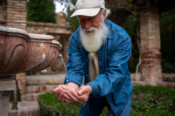 A joyful elderly man with a white beard, wearing a denim jacket and cap, catching water droplets in his hands from a flowing fountain, with a soft smile on his face