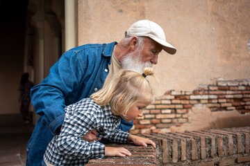 Grandfather and granddaughter leaning over a brick wall, examining something together, with a blurred background of a historical building
