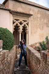 A woman in casual clothing taking photographs in an ancient architectural setting, standing on a cobbled path lined with bricks and greenery, with arches in the background