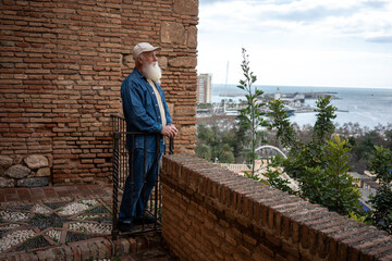 A senior man in a denim jacket leaning on a railing, overlooking the harbor and city skyline, with a scenic view of the coastline and palm trees