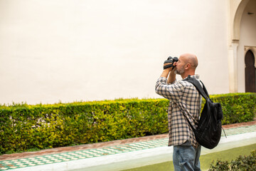 Photographer standing in a garden, taking a photo of a plain white wall with a DSLR camera, wearing a checked shirt and a black backpack