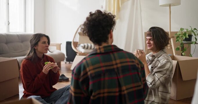 Over the shoulder girl and two guys chat drink tea and eat sandwiches during their lunch break while moving