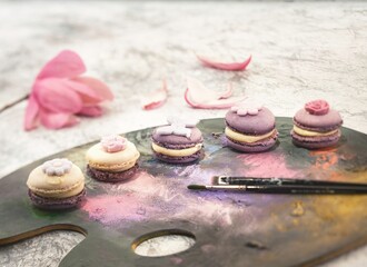 Delicate macarons, adorned with floral decorations, rest on a painter's palette. Petal accents and brushes complete this edible still life scene