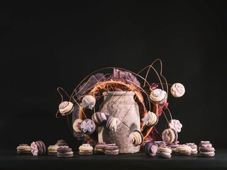 Delicious macarons are scattered around a textured vase, some perched on delicate gold wire branches. The soft light and pastel colors create a whimsical and inviting scene