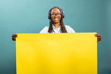 Happy black woman listens to music, grasping blank yellow billboard sign, standing against blue...