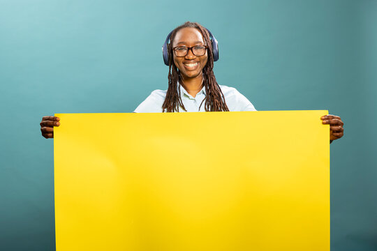 Fototapeta Happy black woman listens to music, grasping blank yellow billboard sign, standing against blue background. Joyful african american female individual holding poster, ready for promotional content.