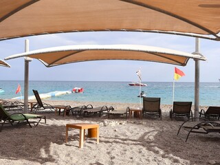 Relaxing beach scene with shade canopies, empty loungers, and a small wooden table on a pebbled shore. Clear blue water, distant boat, and a red-yellow flag create vacation atmosphere.