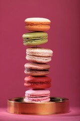 Colorful macarons are stacked on a gold plate in a balanced tower formation. The backdrop is a solid, vibrant shade of pink, emphasizing the dessert presentation