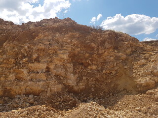 Rugged rock face of a quarry, revealing raw earthy layers and textured stone. A natural, weathered geological backdrop under a summer sky