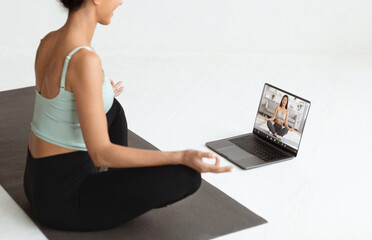 Naklejka premium A young brunette lady sits in lotus position on a fitness mat, engaging in meditation while taking a video call with her yoga instructor.