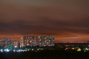 Kyiv cityscape presents a night panorama, featuring modern apartment buildings with many illuminated windows creating a vibrant urban glow against a dark, cloudy sky