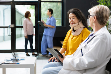 Asian woman with cervical collar attends recovery checkup with medic at hospital lobby, consulting...