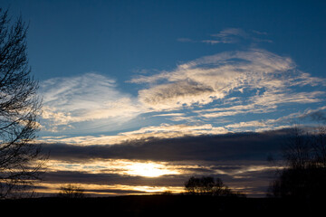 Stunning sunset paints the sky with fiery hues and dramatic cloud layers. Bare tree branches frame this gorgeous evening spectacle. A serene autumn skyscape