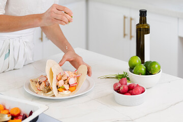 Woman squeezing fresh lime juice over delicious fish tacos with radishes and carrots on white kitchen counter