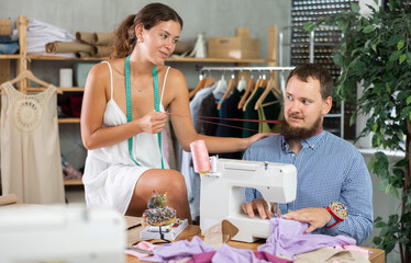 Playful young woman harasses male tailor working at a sewing machine in fashion studio