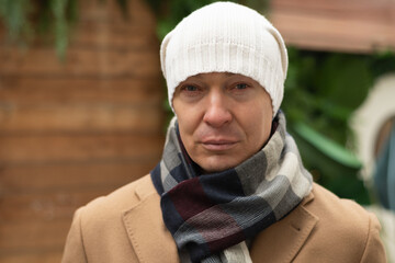 Portrait of serious adult caucasian handsome man wearing white beanie thinking with blurred Christmas background at winter street. Cold weather.