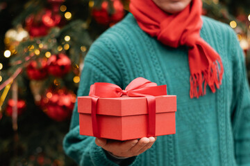 Christmas red gift box with red bow in man hand against Christmas tree. Xmas giving and shopping. Boxing Day. Close up.