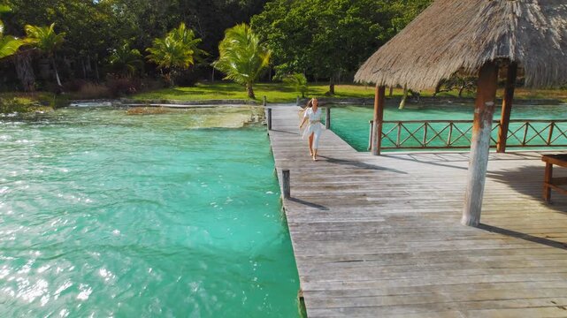 Woman walking on wooden pier by turquoise water under tropical hut in Mexico