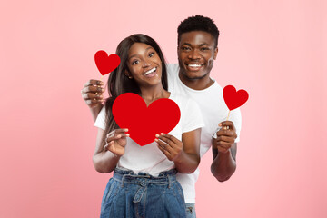 A young romantic black couple poses happily while holding red heart cards. They are smiling and enjoying the special moment together, capturing the essence of love on a playful pink background.