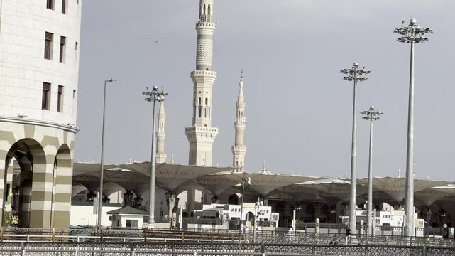 Iconic view of the towering minaret of the Prophet's Mosque (Al-Masjid an-Nabawi) against a bright sky, surrounded by palm-lined trees and city life in Medina. Use for travel, religious documentaries.