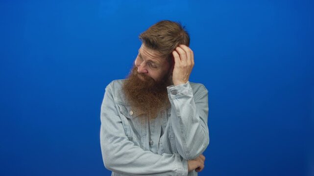 Man scratches head in studio wearing a thick beard and denim jacket with arms crossed while tilting sideways; thinking.