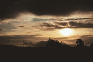 A dramatic sunset sky with warm golden sunlight breaking through layered clouds, casting a soft glow over a rural horizon. The silhouettes of trees and power poles add depth and contrast to the scene,