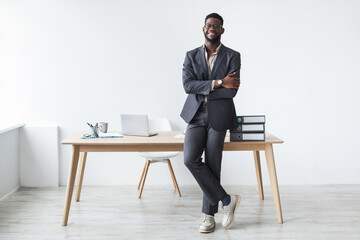 A cheerful young black businessman in a formal suit stands with crossed arms near his office desk....