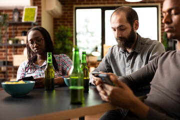 Caucasian male individual uses cellphone to browse online, with beer bottle placed on table in front of him. Bearded white man with mobile device, sits in middle of friends, enjoying their company.