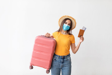 A young woman in a yellow shirt and straw hat holds a pink suitcase and travel documents, smiling and ready for her journey. She wears a protective mask for safety.