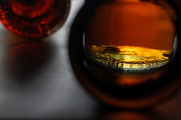 Rich amber liquid reflecting light on glass bottom of bottle, viewed through the dark, circular neck of another bottle, suggesting moody and dramatic atmosphere