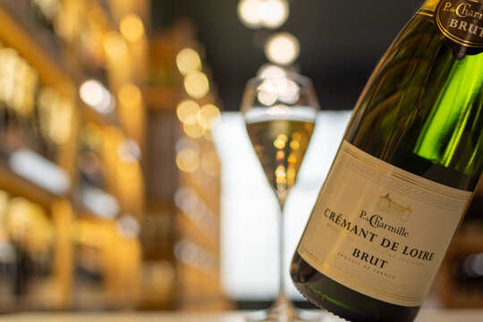 Bottle of P.G. Charmille Cr&eacute;mant de Loire Brut against luminous full glass of sparkling wine and warm golden-hued blurred shelving of wine shop in background. Ukraine, Zhytomyr, October, 29, 2025