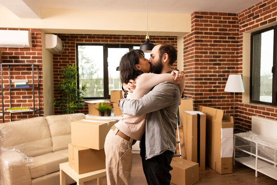 Caucasian couple kisses and embraces in living room after purchasing apartment, excited to move in together. Carefree husband and wife share tender moment surrounded by cardboard boxes in new home.