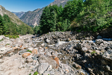A rocky mountain riverbed winds through a lush green valley with towering peaks under a clear blue sky on a bright sunny day.