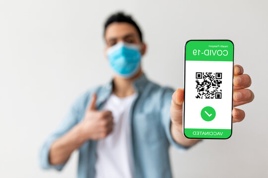 A man wearing a blue mask shows his smartphone proudly. The screen displays a vaccination status with a QR code. He gives a thumbs up in a well-lit indoor space.