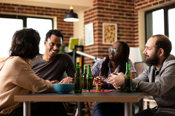Small gathering of young adults engaged in conversation and playing indoor game around table. Diverse group takes break from board game, with everyone attentively listening to black man share a story.