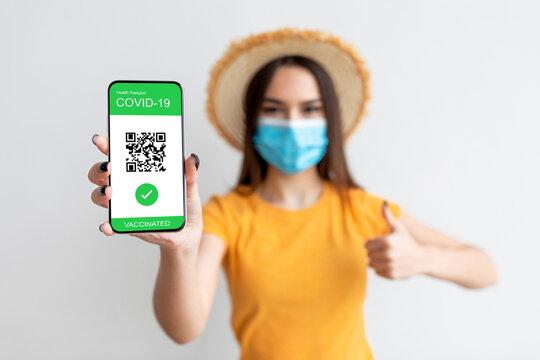 A young woman, wearing a yellow shirt and a straw hat, holds her smartphone displaying her COVID-19 vaccination passport. She gives a thumbs up, showing her approval.