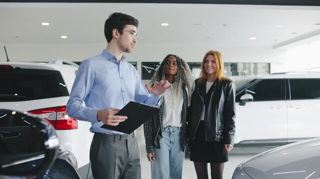Two women, a lesbian couple, explore a car dealership with a male salesperson, learning about vehicle options and enjoying the experience together