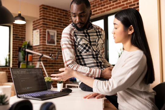 Multiethnic couple collaborating in home office, one seated and one standing, reviewing programming content on laptop. Warm lighting, teamwork and diverse partnership in brick wall apartment.