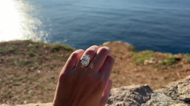 Elegant close up of a woman&rsquo;s oval diamond engagement ring catching the sunlight against the rocky shore of the Croatian coastline in landscape format.