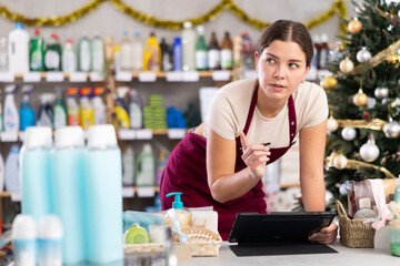 Young woman seller conducting inventory with tablet in household chemicals store decorated for christmas