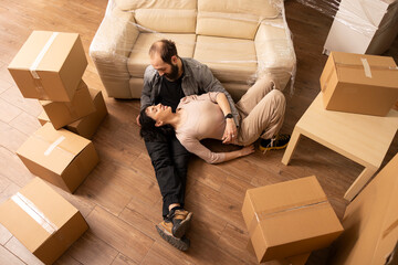 Bearded man resting on floor with beautiful woman, surrounded by cardboard boxes, communicating about furniture and home decorations. Happy relationship moments of caucasian homeowners on moving day.