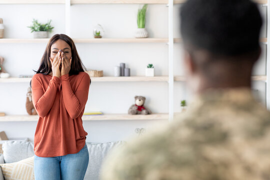 A woman expresses surprise and joy while meeting a soldier in a welcoming living room filled with plants and decorations, highlighting their emotional connection.