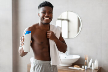 A cheerful African American man stands in a modern bathroom, smiling at the camera. He holds a...