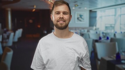Man cupping hand to ear listening in building dining area wearing white t shirt and beard; curiosity listening engagement.