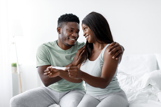 A joyful couple smiles and embraces while holding a pregnancy test in a bright and comfortable bedroom. Their expressions show happiness and anticipation for their future.